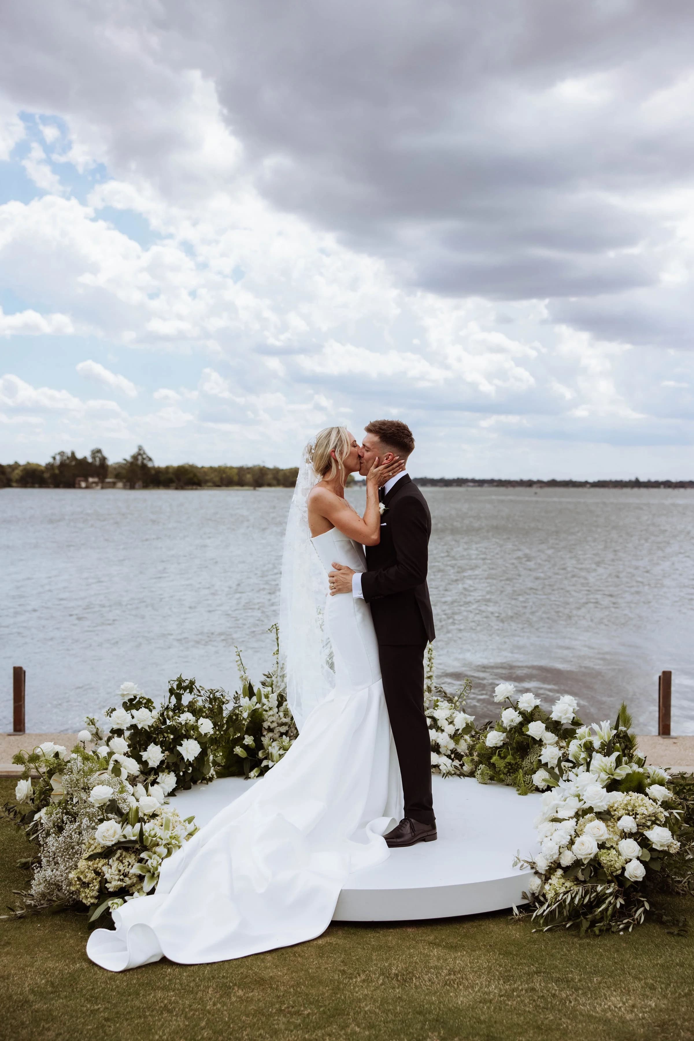 Ben and Tempe kissing on a round white platform ringed with white flowers, beside a lake under a cloudy sky.