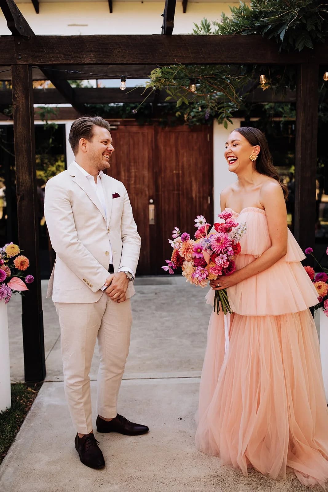 Maddy and Andrew laughing together under a timber pergola on their wedding day.